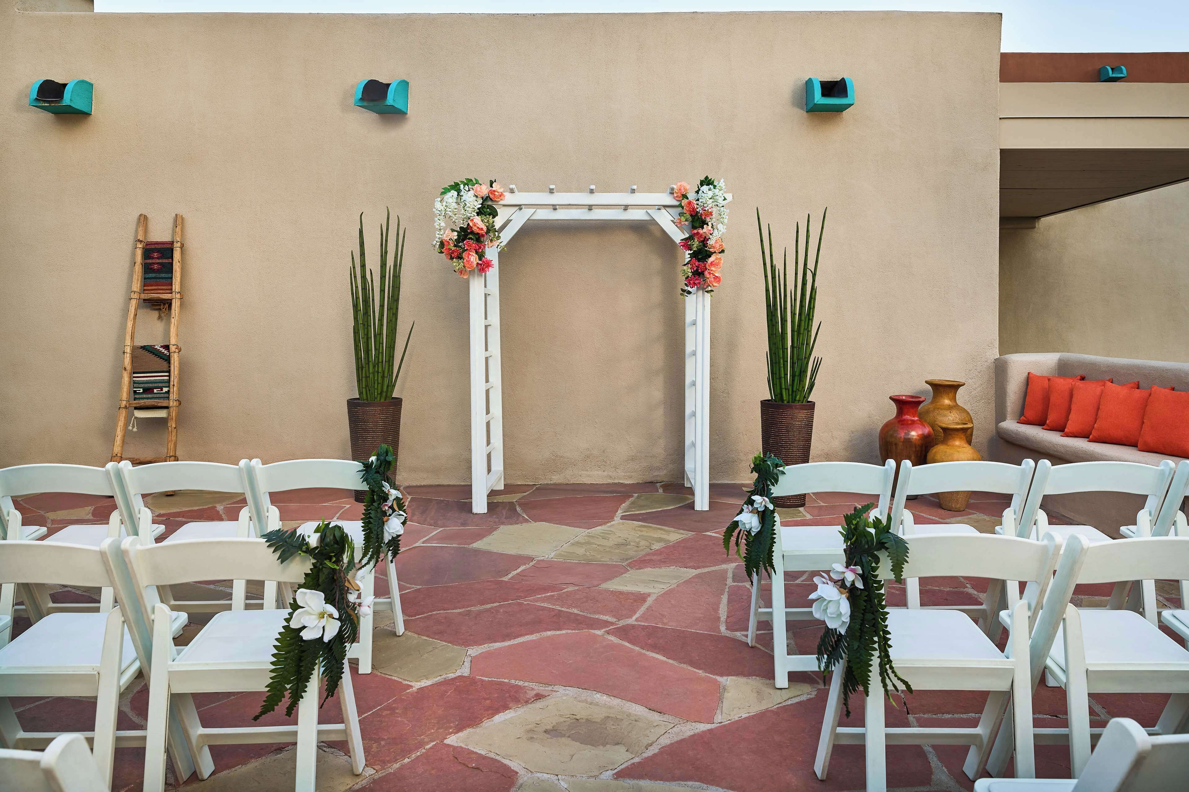 Un petit espace extérieur avec des chaises blanches disposées en demi-cercle, une arche blanche ornée de fleurs et des plantes en pot.