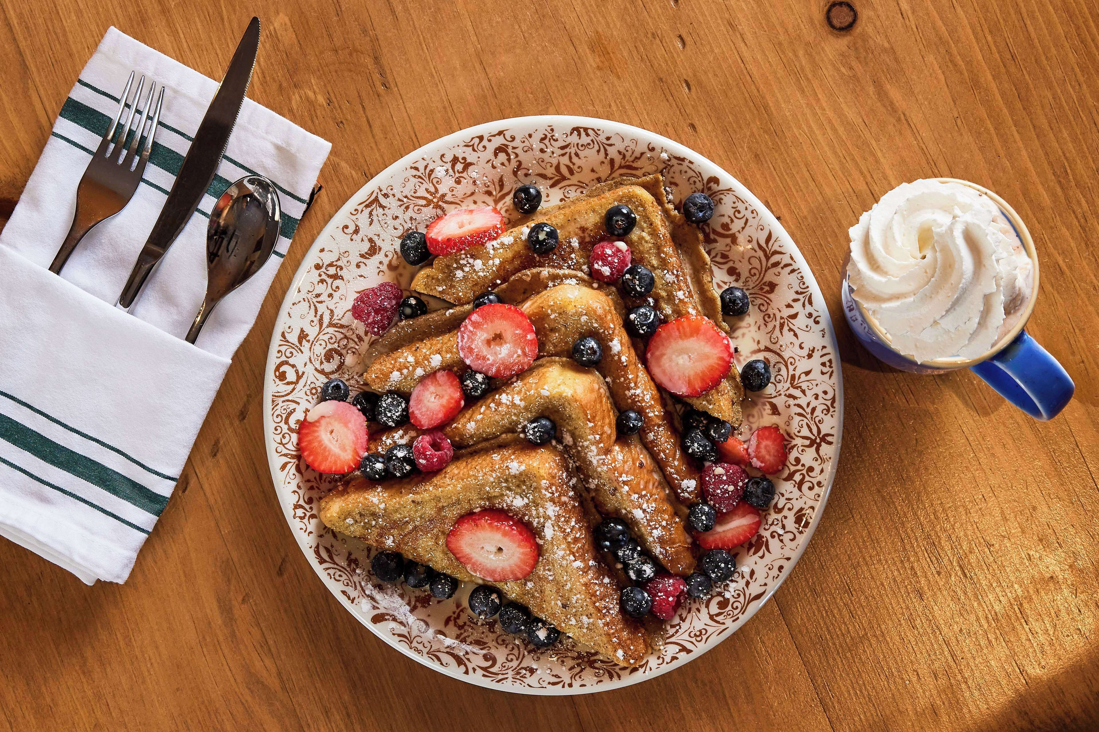 A plate of French toast with strawberries and blueberries, a fork and knife on a napkin, and a cup of whipped cream.