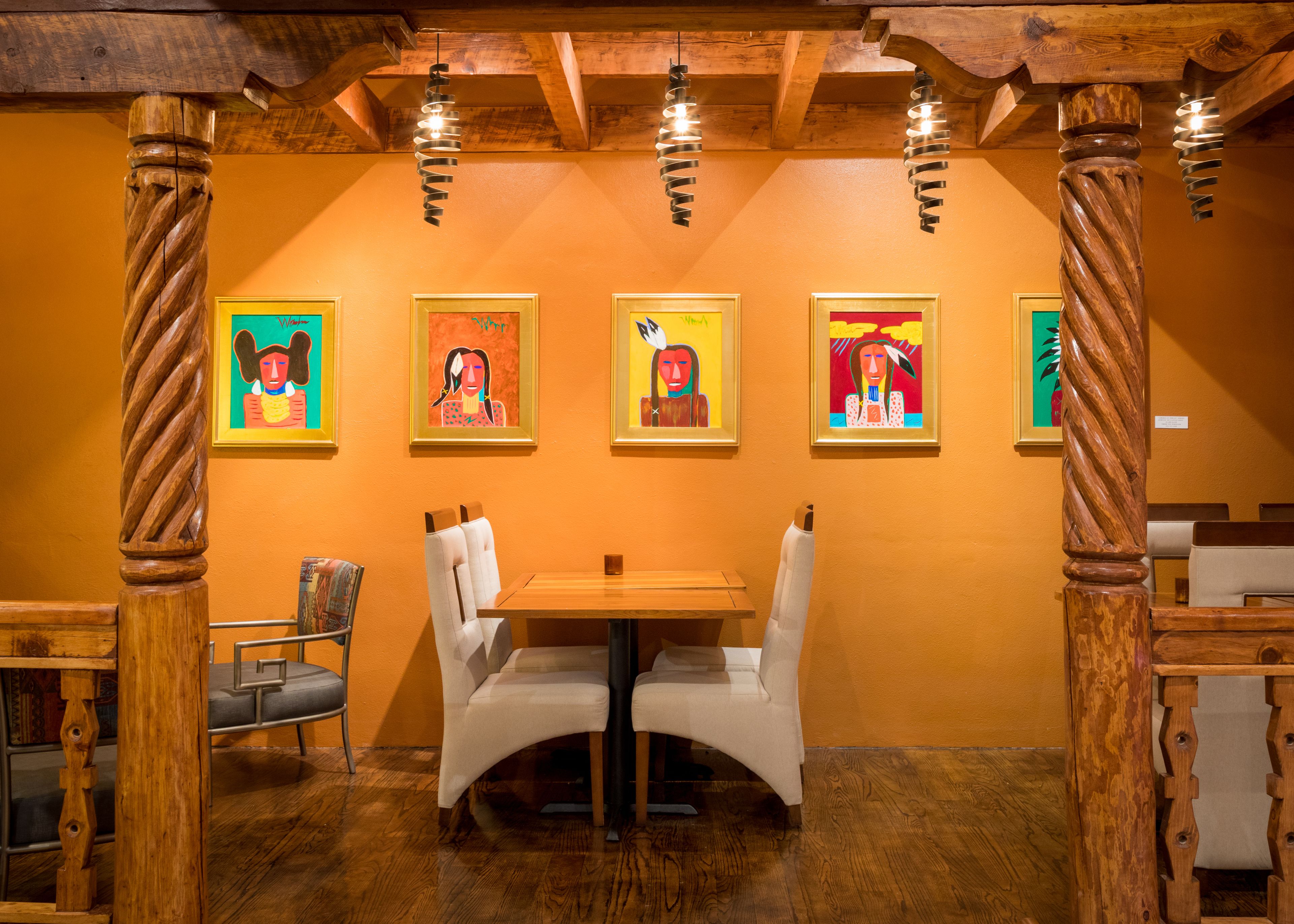 Uma sala de jantar com vigas de madeira, uma mesa de madeira, cadeiras estofadas brancas e obras de arte emolduradas na parede.