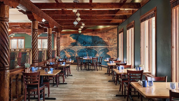 Un restaurante con mesas y sillas de madera, un mural en la pared y ventanas amplias.