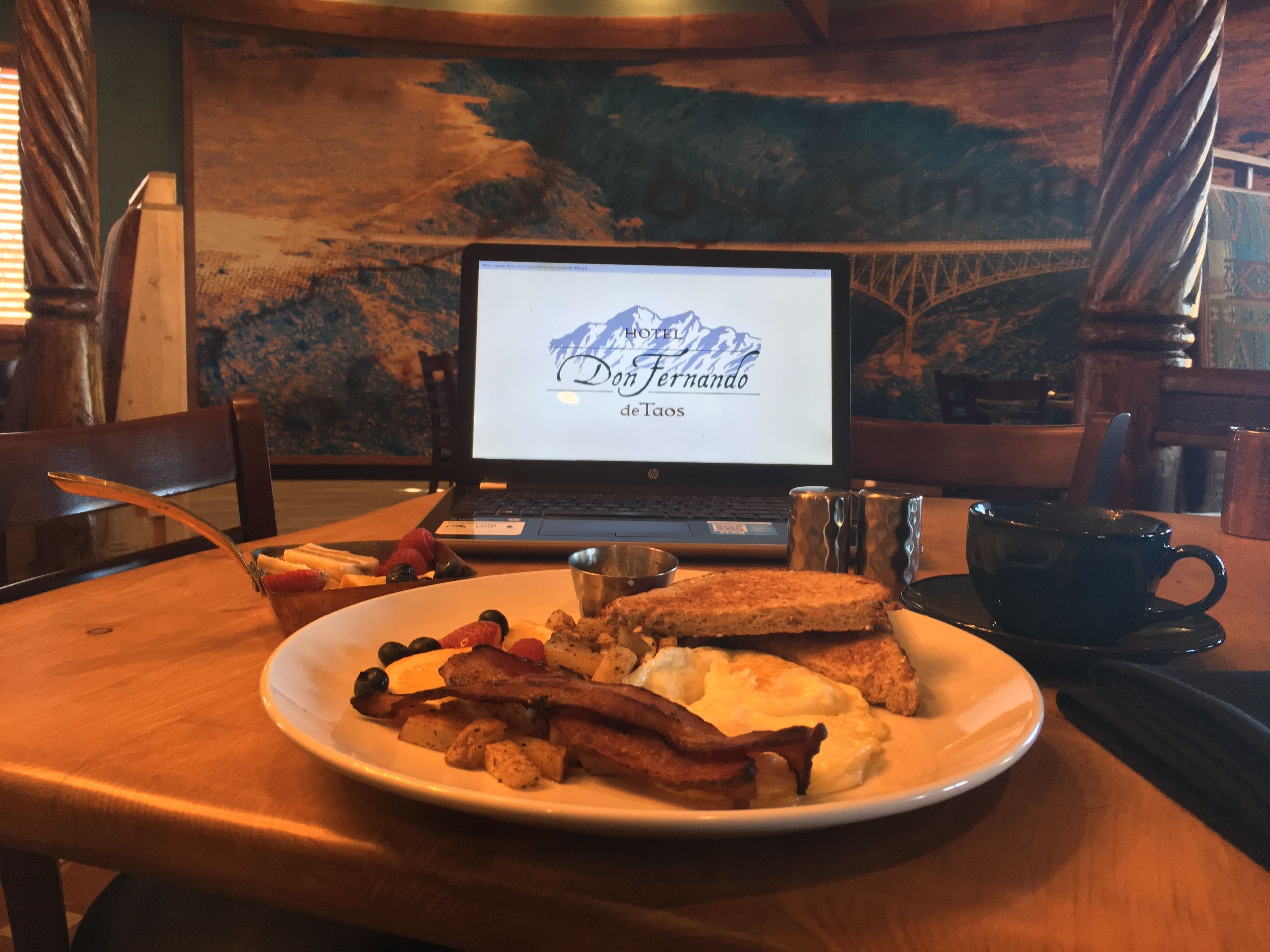 A breakfast plate with bacon, eggs, toast, and berries on a wooden table. A laptop displaying 'Dom Fernando de Taos' is in the background.