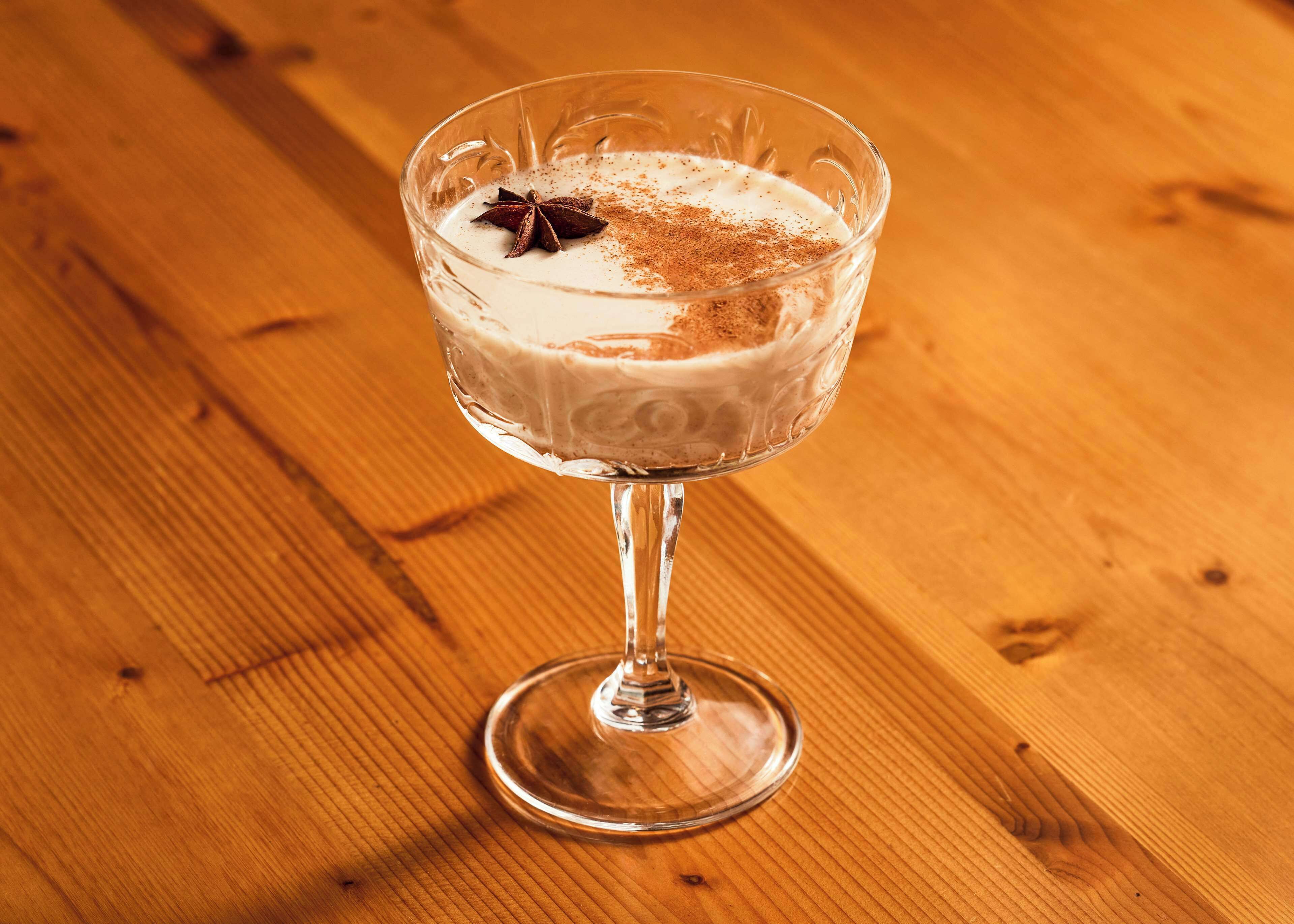 A clear glass with a creamy beverage, a star anise, and a sprinkle of spice on a wooden surface.