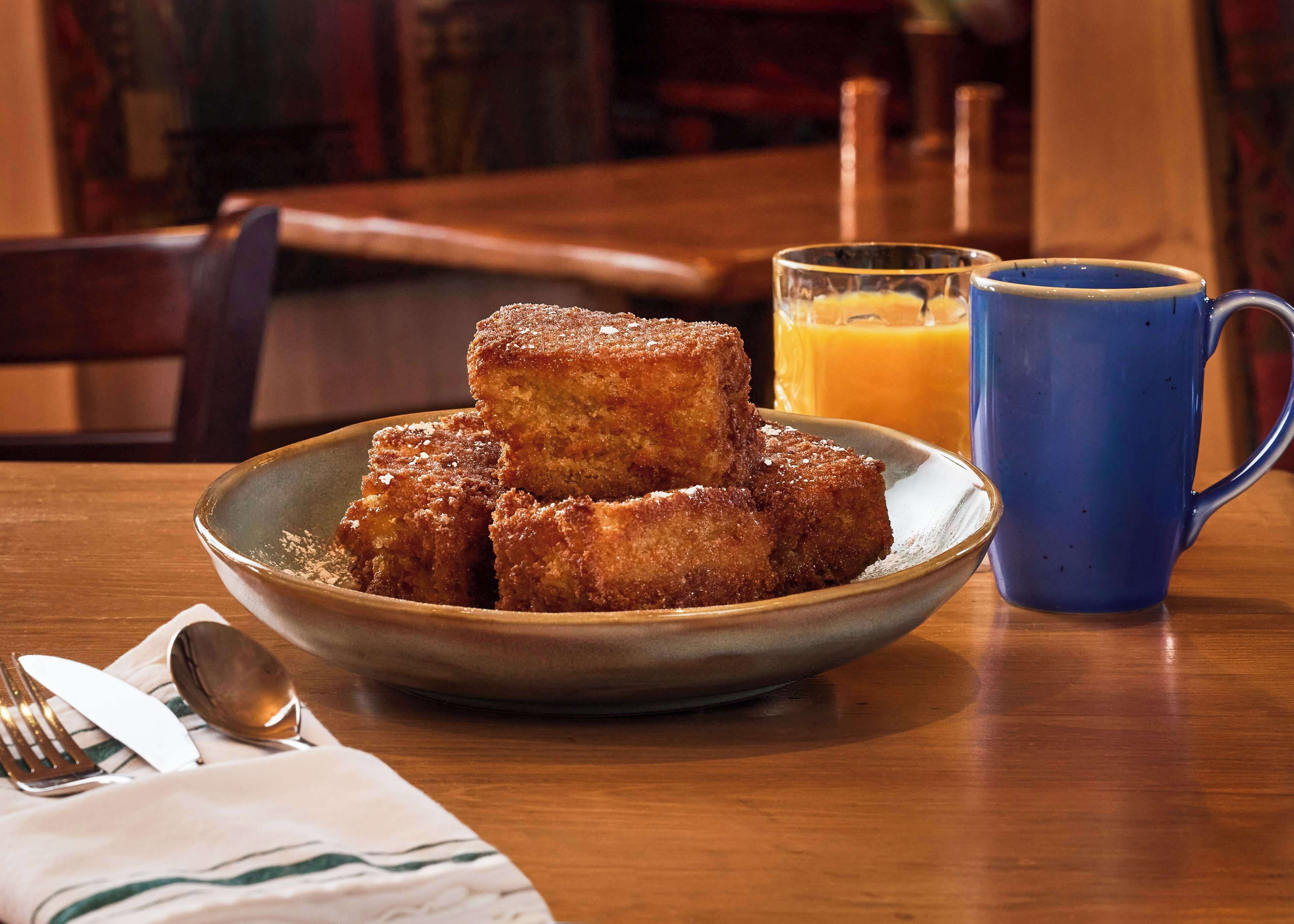 Une assiette de pain perdu nappé de sirop, une tasse bleue et un verre de jus d’orange sur une table en bois.