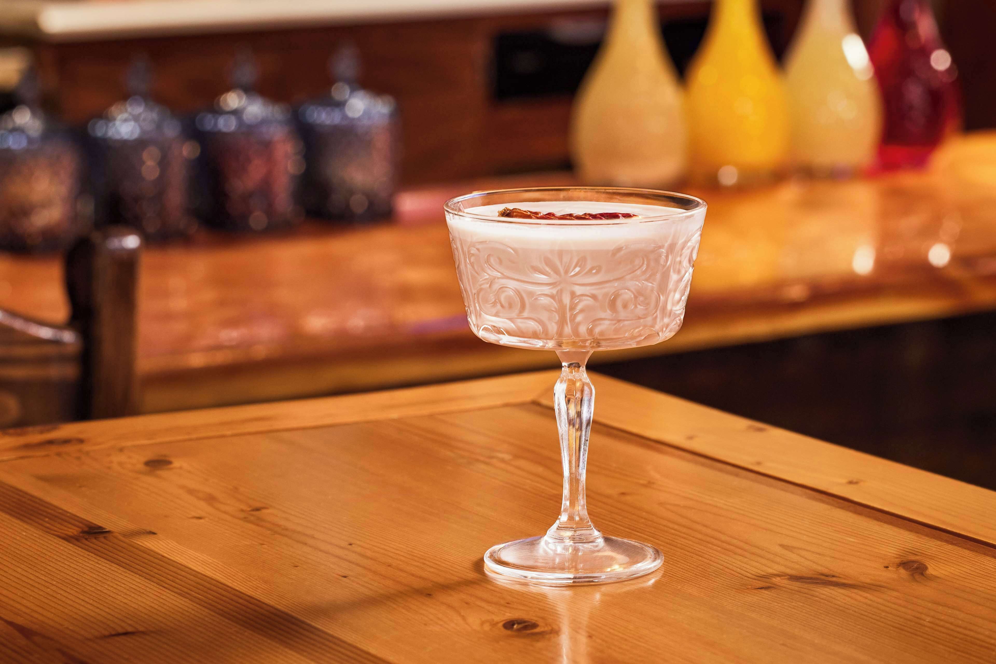 Cóctel transparente en un vaso sobre una mesa de madera, con un fondo desenfocado de utensilios de bar.