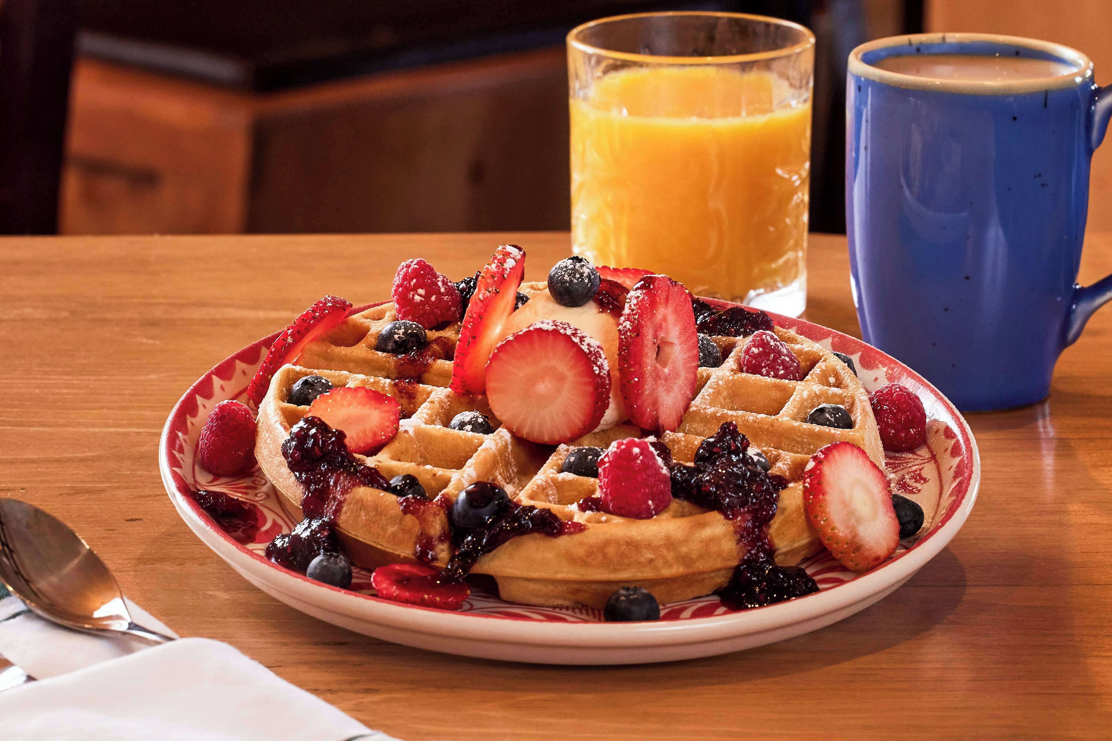 A plate of waffles topped with strawberries and blueberries, a glass of orange juice, and a blue mug on a wooden table.