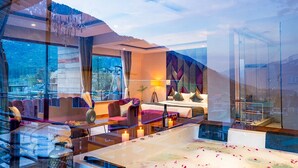 A modern hotel room with a large bed, a sitting area with a sofa and armchairs, a glass coffee table, and a view of the mountains. -  Moustache Manali (Manali)
