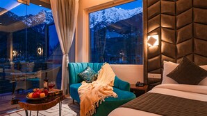 A modern hotel room with a large window offering a mountain view, a teal sofa with a throw blanket, a wooden side table with fruit and wine, and a bed with a quilted headboard. -  Moustache Manali (Manali)