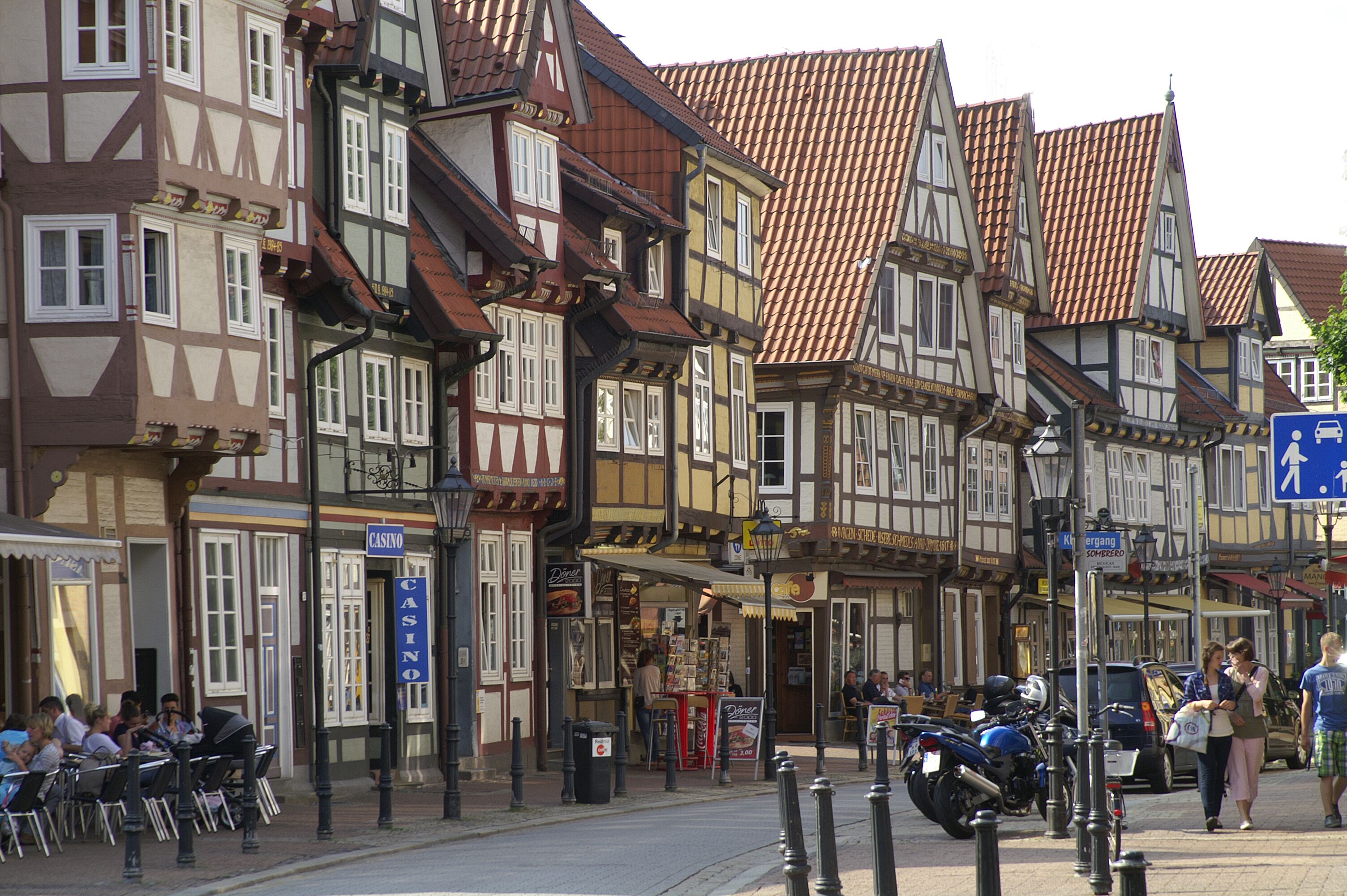 Una calle europea tradicional con edificios de entramado de madera, mesas al aire libre en un café y un cartel de casino a la vista.