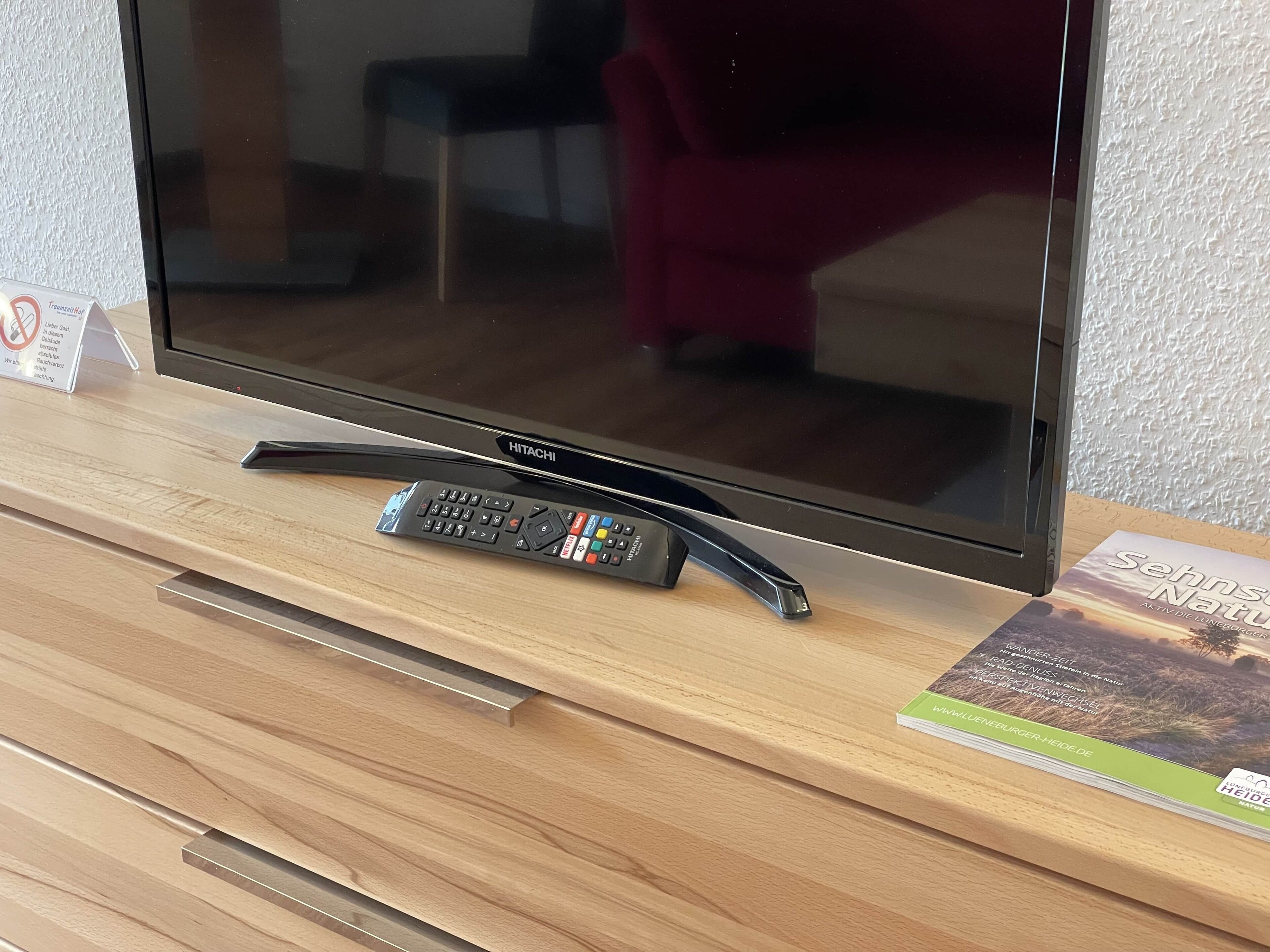 Ein Fernseher auf einer Holzkommode, neben ihm eine Fernbedienung und ein Buch.