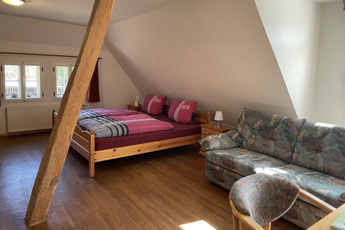 A bedroom with a sloped ceiling, a bed with a patterned bedspread, a wooden nightstand, a patterned sofa, and a window with curtains.