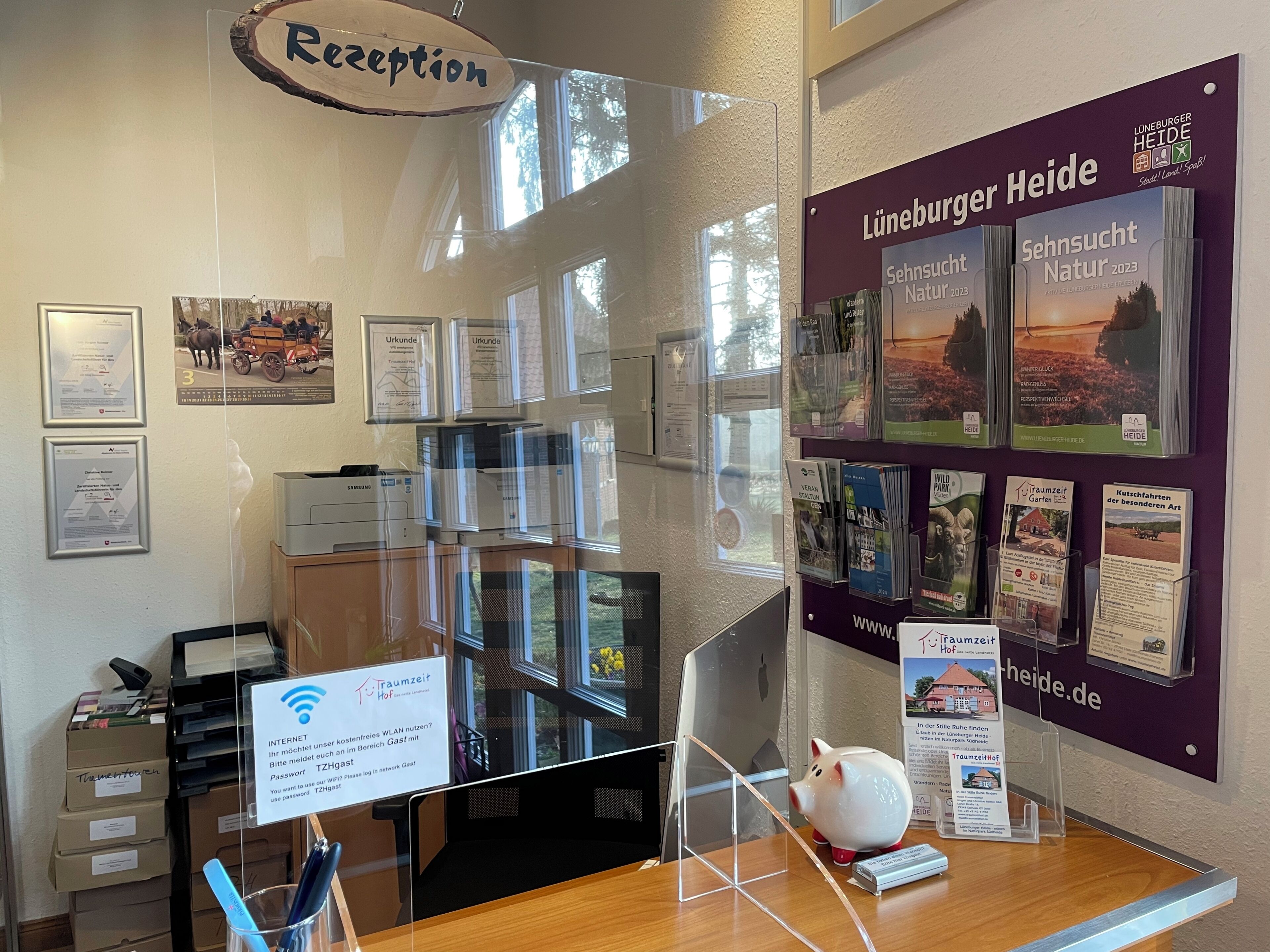 Ein Empfangstresen mit einem Schild mit der Aufschrift 'Reception', einem Computer, einem Drucker und verschiedenen Informationsplakaten an der Wand.
