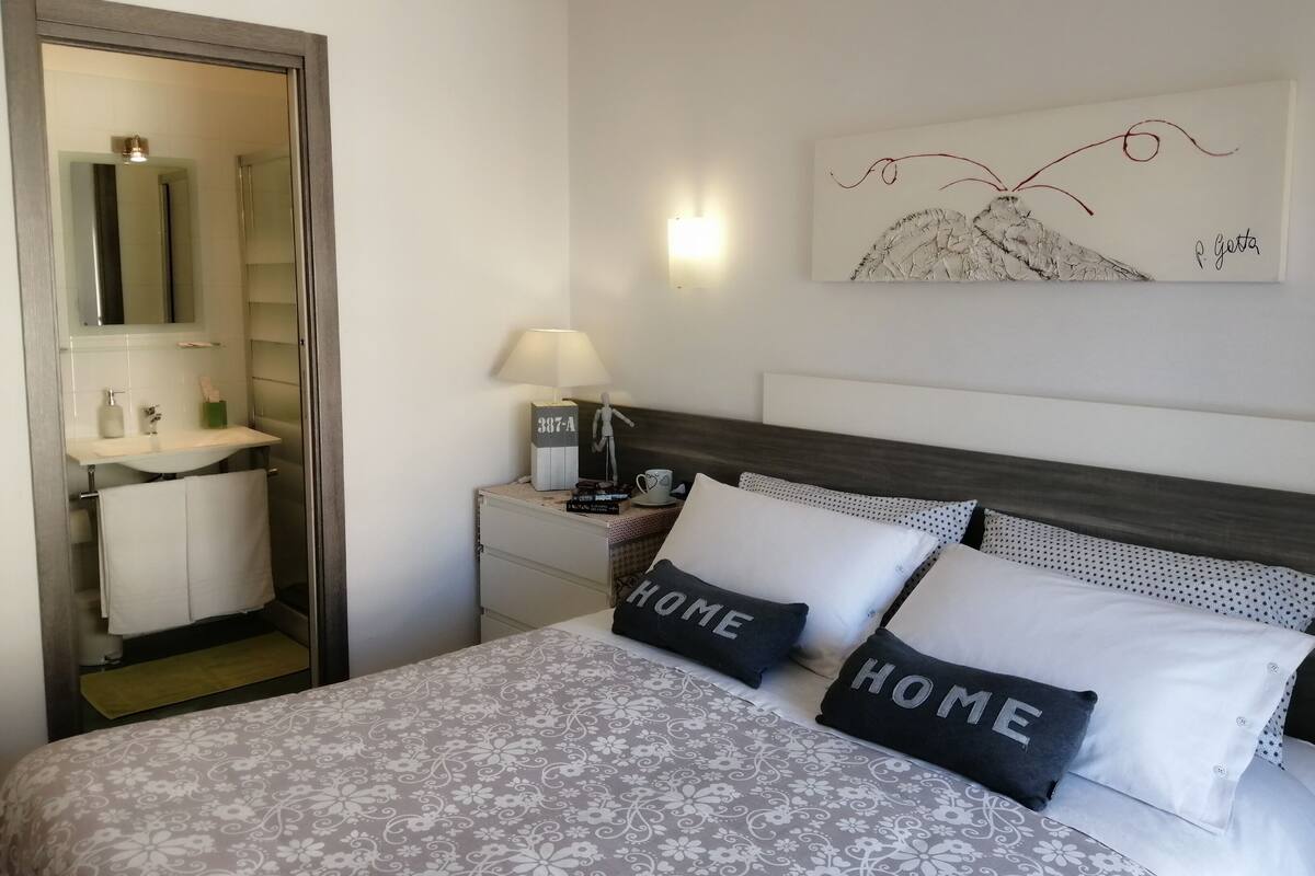 Une chambre à coucher avec un lit, deux oreillers ornés du mot « HOME », une table de chevet avec une lampe, une œuvre d’art accrochée au mur et une salle de bain visible à travers une porte ouverte.