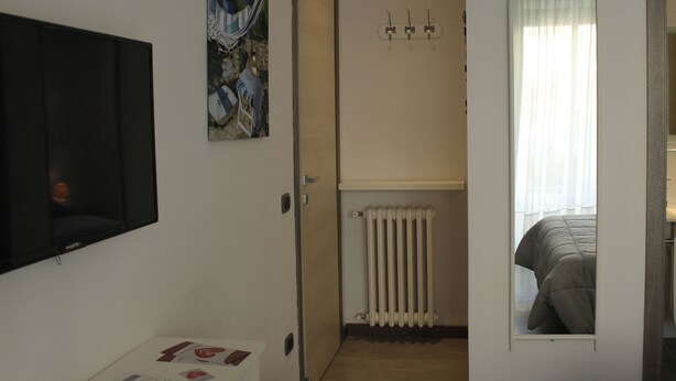 Ein Schlafzimmer mit an der Wand montiertem Fernseher, einem Gemälde, einem Heizkörper und einem Spiegel, der ein Bett reflektiert.