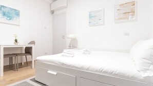 Une chambre à coucher moderne et minimaliste, avec un lit blanc, un bureau et deux chaises. Trois œuvres d’art encadrées ornent le mur.