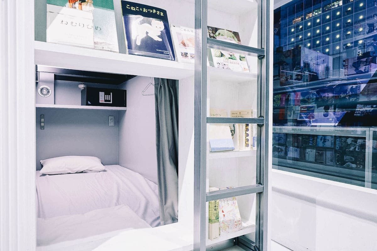 A compact room with a bed, a small desk with a computer, and a glass wall displaying a cityscape.