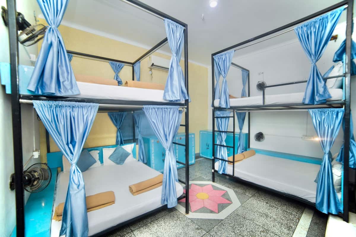 A dormitory room with bunk beds, each with a curtain. There are storage boxes and a fan on the floor.