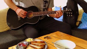 Iemand speelt op een akoestische gitaar op een bank, met een bord met toast en jam, en een kom soep op een houten tafel.