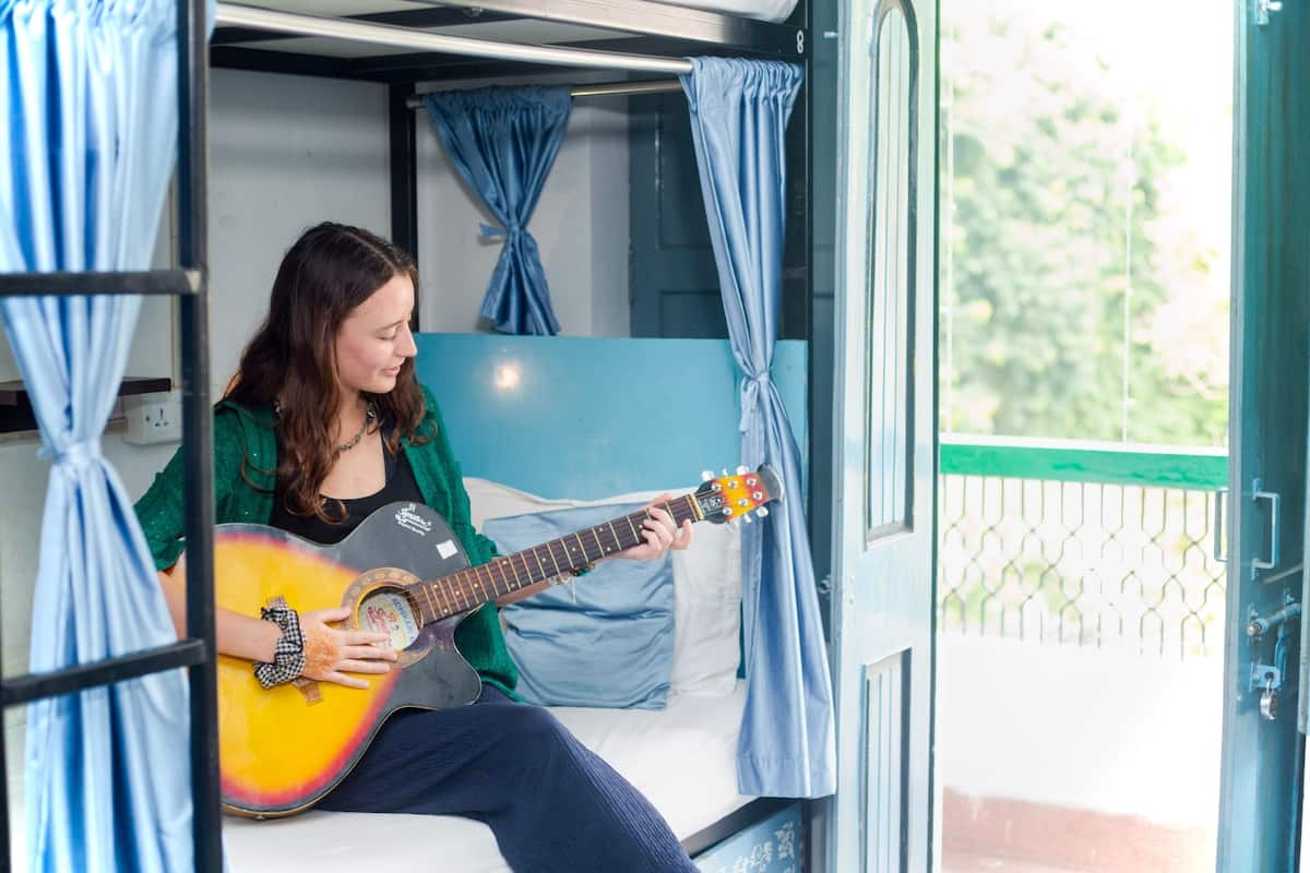 A person sitting on a bed in a room with a window, playing a guitar.