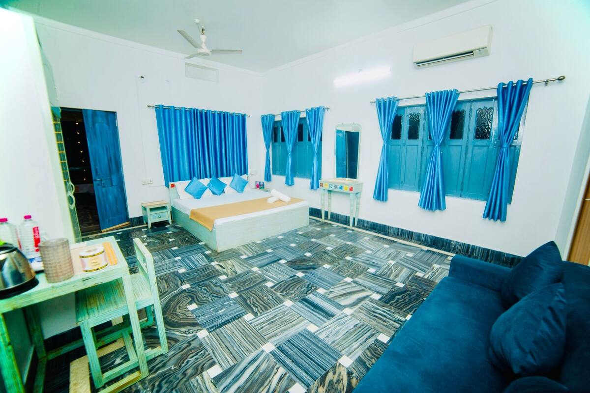 A bedroom with a bed, a desk, and a chair. There is a window with curtains, a ceiling fan, and an air conditioning unit.
