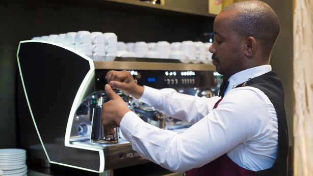 Een barista maakt koffie in een professionele omgeving, met een koffieapparaat en stapels wegwerppapieren bekers.