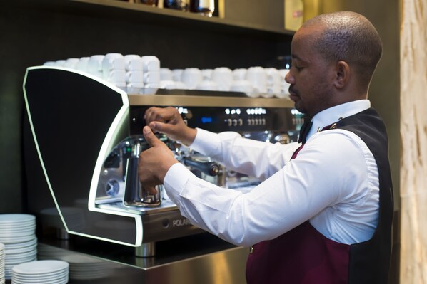 A barista preparing coffee in a commercial setting with a coffee machine and stacks of disposable cups. - Doubletree by Hilton Pointe-Noire (Pointe-Noire)
