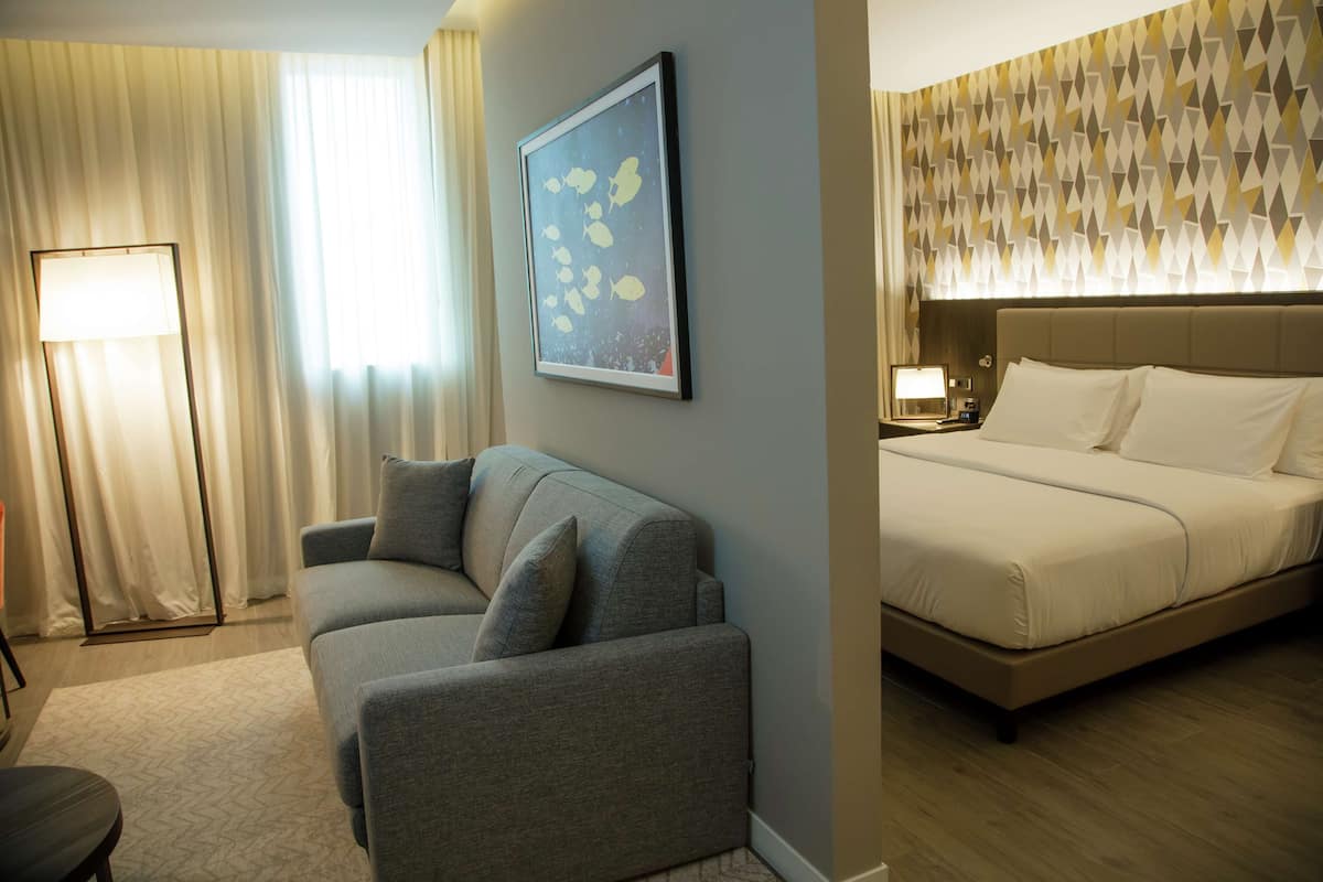Ein Hotelzimmer mit grauem Sofa, einem Bett mit weißer Bettwäsche, einem gerahmten Bild an der Wand und einer Wandtapete mit Muster.