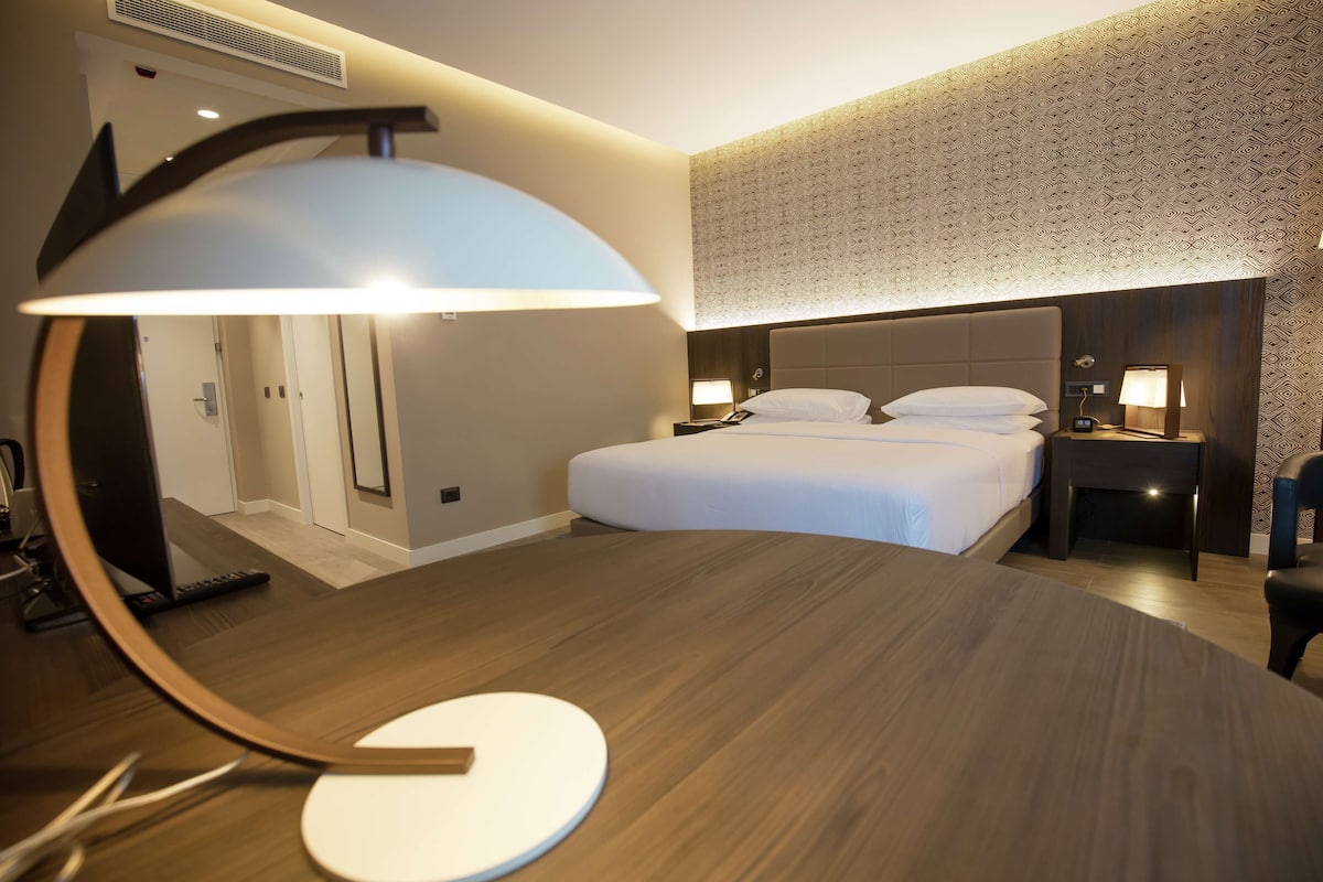 Ein Hotelzimmer mit Bett, Nachttischen, einem Schreibtisch mit Lampe und einem Spiegel.