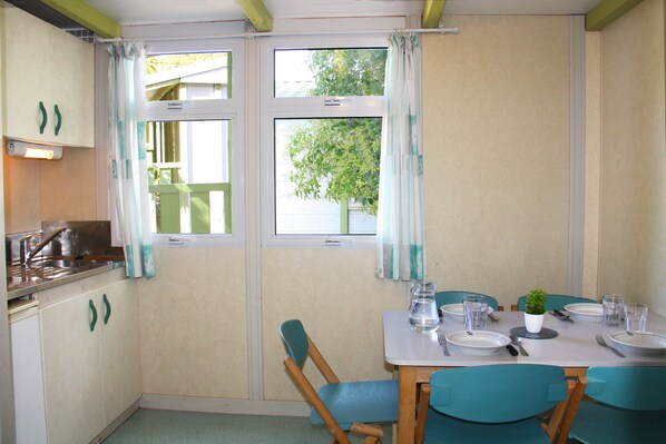 A dining area with a table set for four, chairs, a window with curtains, and a kitchen area with a sink and cabinets. - Village Vacances Azureva Sigean (Sigean)
