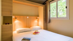 A neatly made bed with a hat and sunglasses on the side, a window with a view of trees, and a shelf with two items. - Village Vacances Azureva Sigean (Sigean)