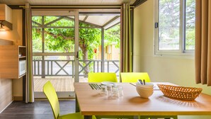 A dining area with a wooden table, green chairs, and a view of a wooden deck and trees. - Village Vacances Azureva Sigean (Sigean)