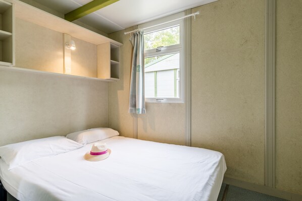 A single bed with white bedding, a hat on the pillow, and a window with a curtain. - Village Vacances Azureva Sigean (Sigean)