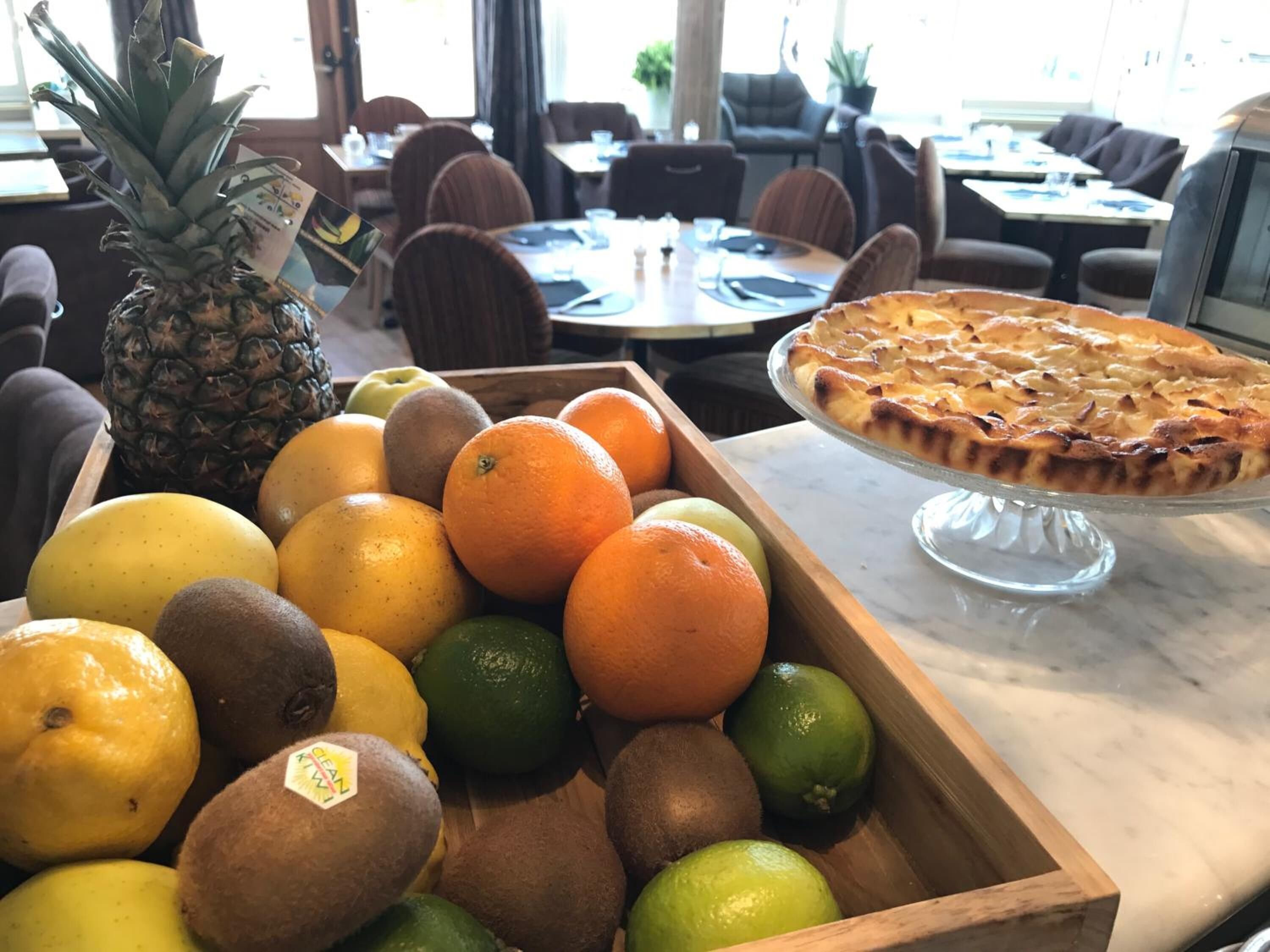 Een houten fruitschaal met ananas, sinaasappels, kippenfruit en limoenen, plus een taart op een glazen presentatiebord in een restaurant.