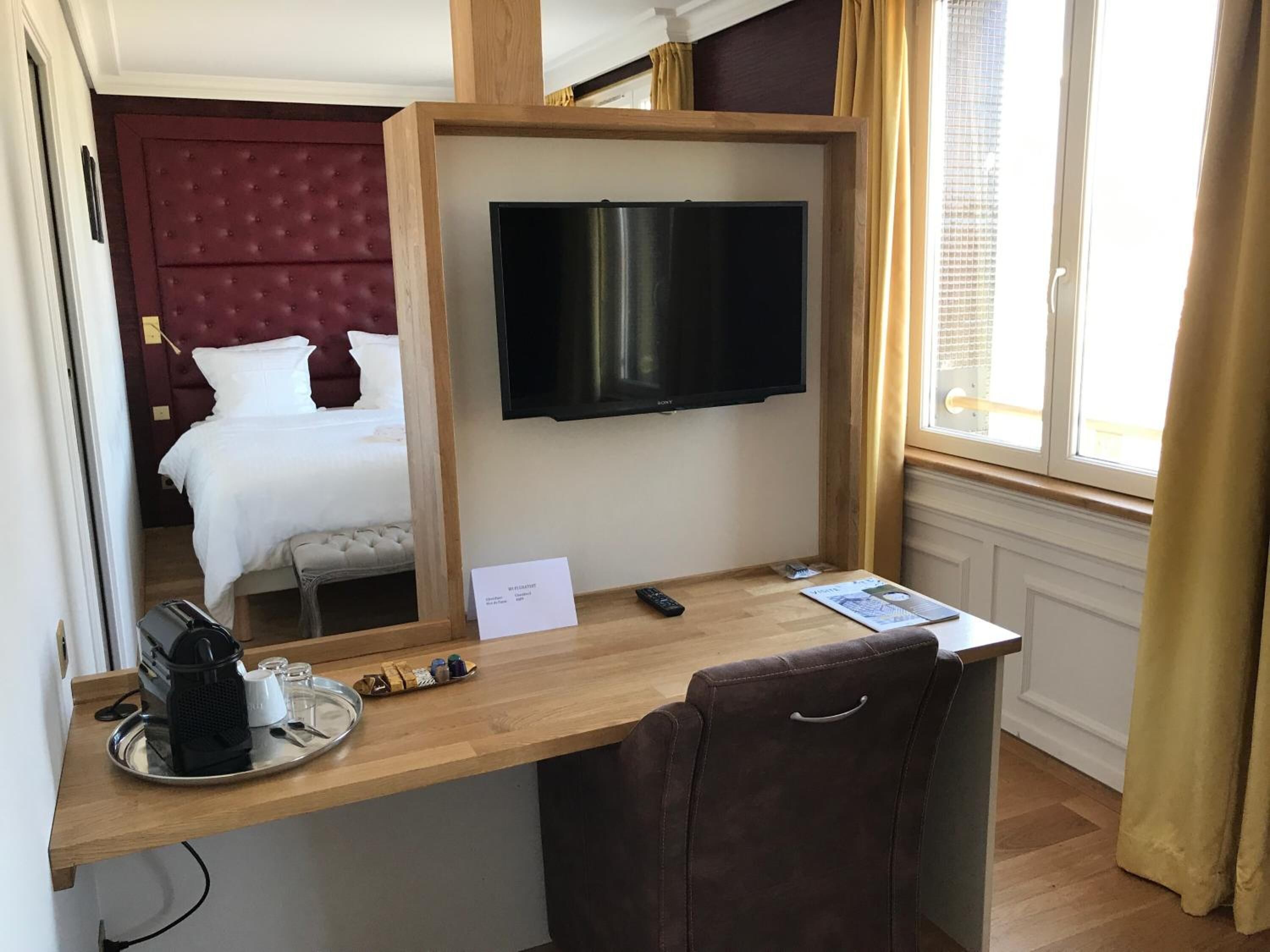 A hotel room with a bed, a TV mounted on a wooden frame, a desk with a chair, and a window with curtains.