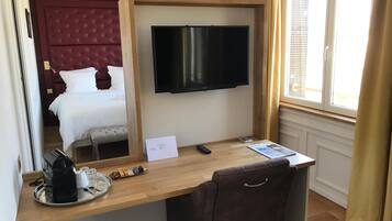 A hotel room with a bed, a TV mounted on a wooden frame, a desk with a chair, and a window with curtains.