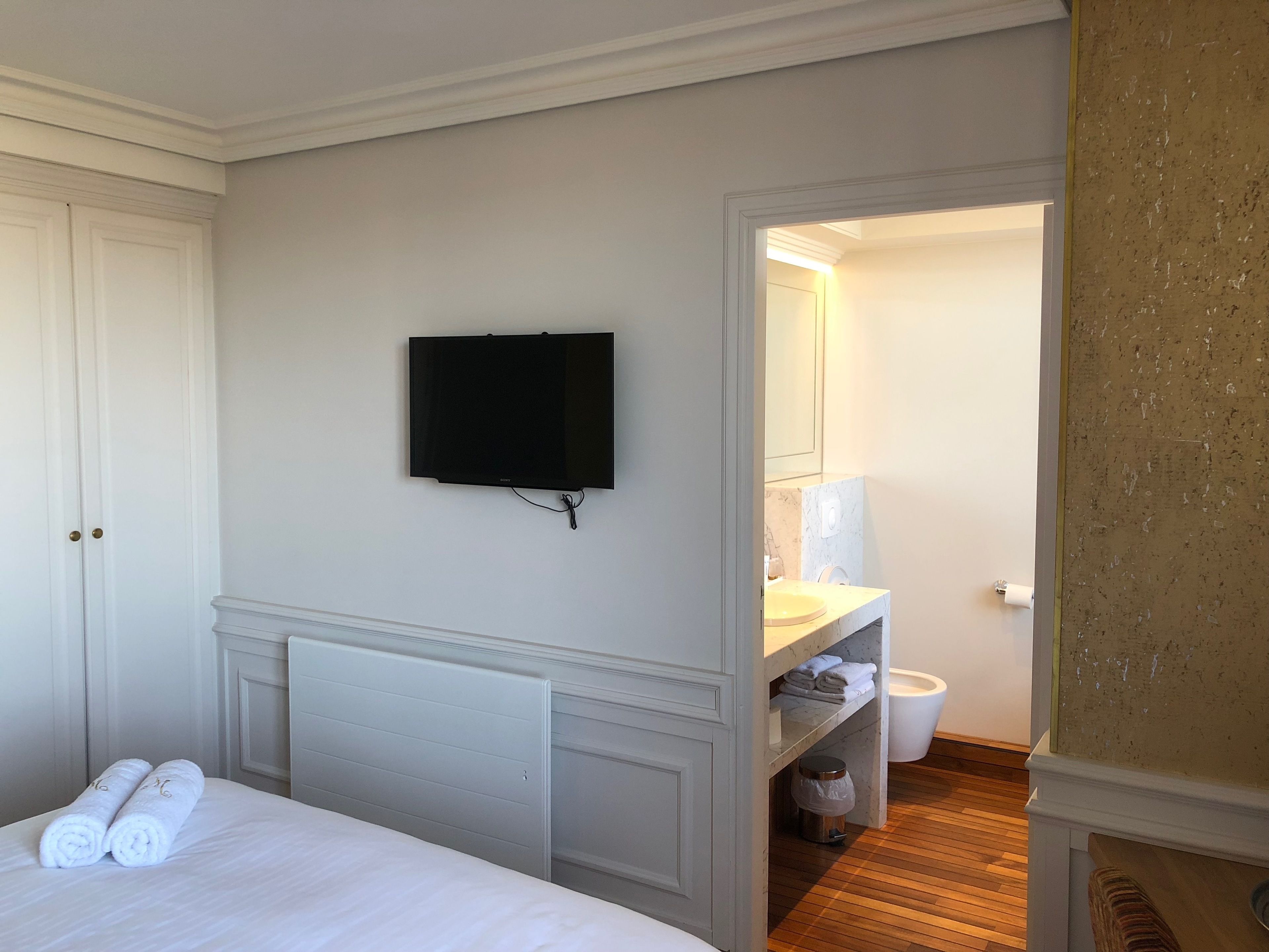 Une chambre d’hôtel comprenant un lit, un téléviseur à écran plat fixé au mur, une salle de bain avec un lavabo et un miroir, et un plancher en bois.