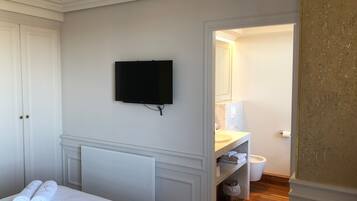 Une chambre d’hôtel comprenant un lit, un téléviseur à écran plat fixé au mur, une salle de bain avec un lavabo et un miroir, et un plancher en bois.