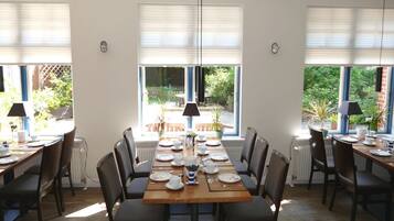 Un coin repas avec une longue table dressée pour six personnes, encadré de deux fenêtres équipées de stores, laissant entrer la lumière naturelle.