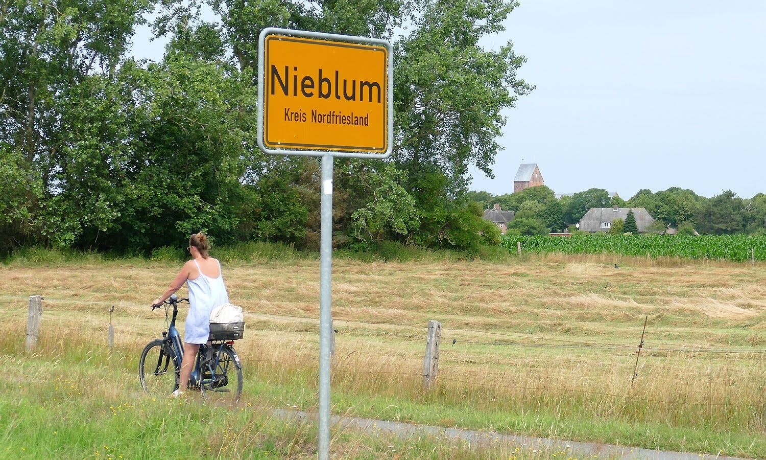 A sign reading "Niebum Kreis Nordfriesland" in a rural setting with a cyclist and a field.
