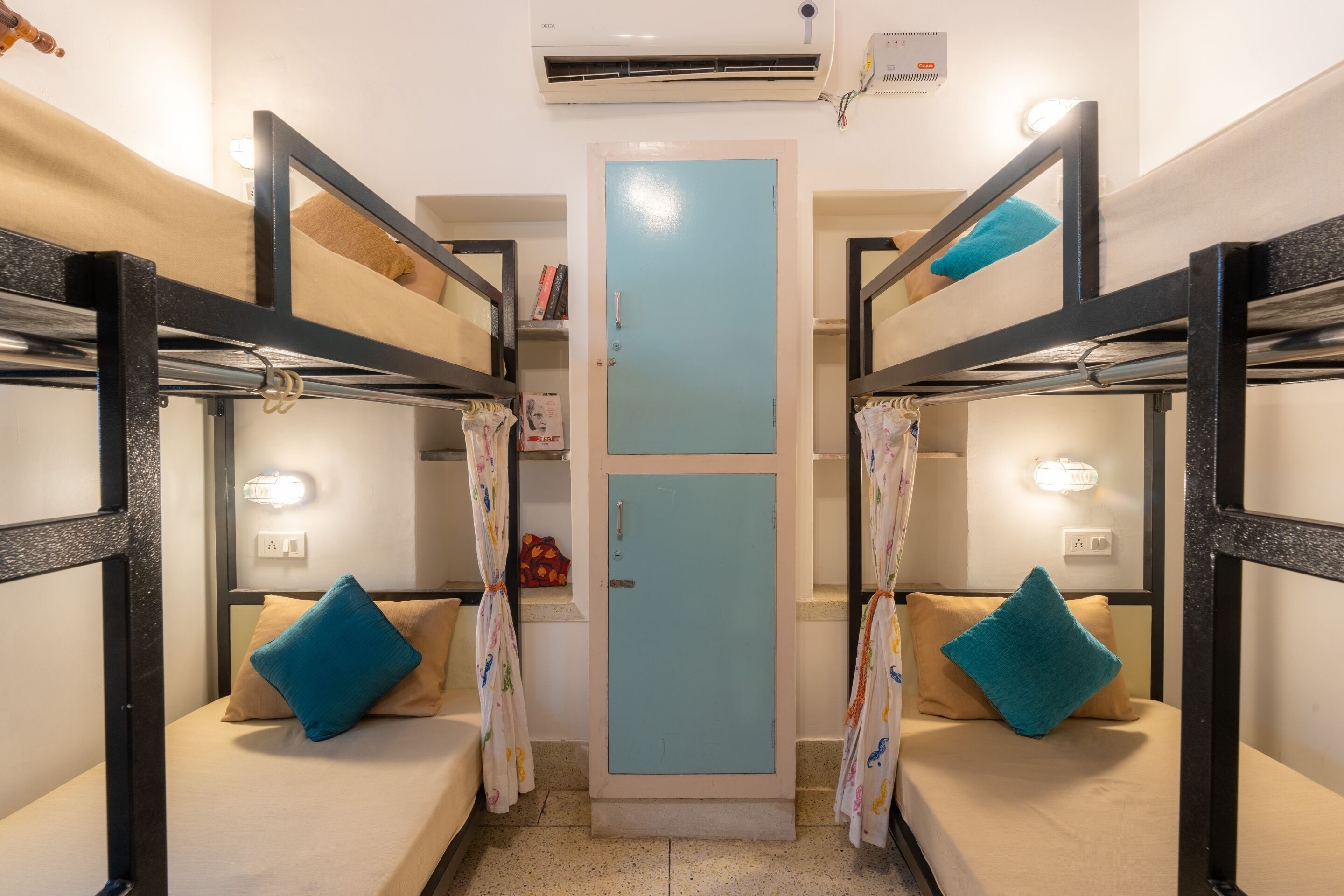 Habitación con literas, una puerta central, almohadas en cada litera y un estante pequeño con libros.