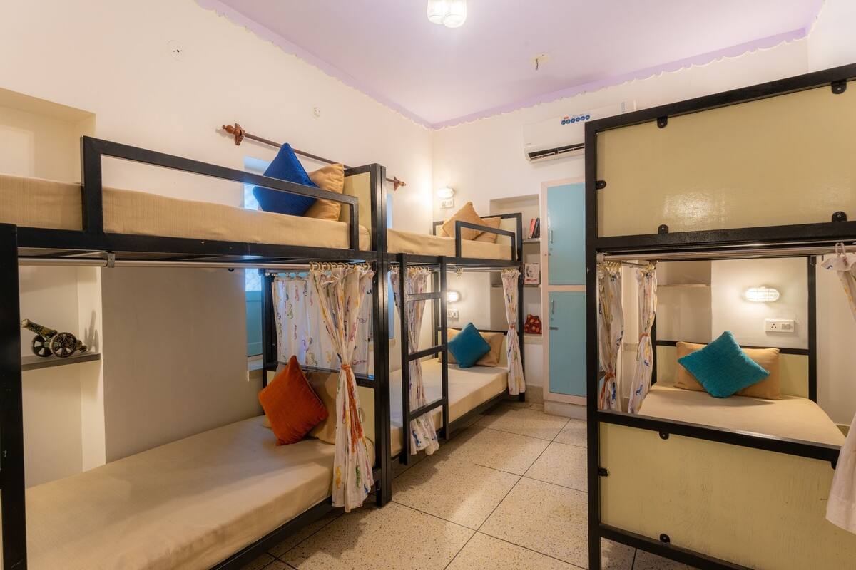 Une chambre de dortoir avec des lits superposés, chacun étant muni d’un rideau. Il y a des oreillers sur les lits et une étagère avec des objets décoratifs.