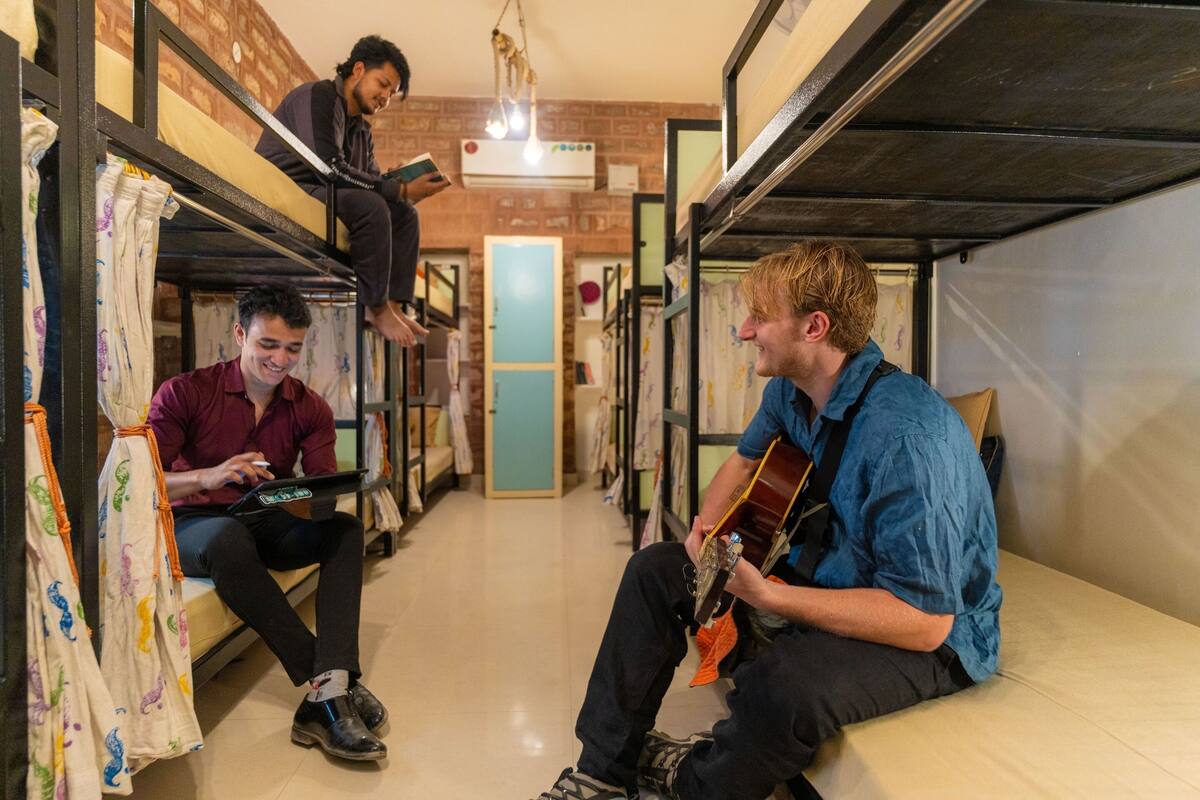 Une chambre d’auberge de jeunesse avec des lits superposés, une personne qui joue de la guitare et une autre qui utilise un ordinateur portable.