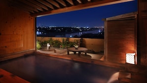Un onsen japonais traditionnel, avec un intérieur en bois, un bain d’eau chaude et une vue sur le paysage urbain illuminé la nuit.