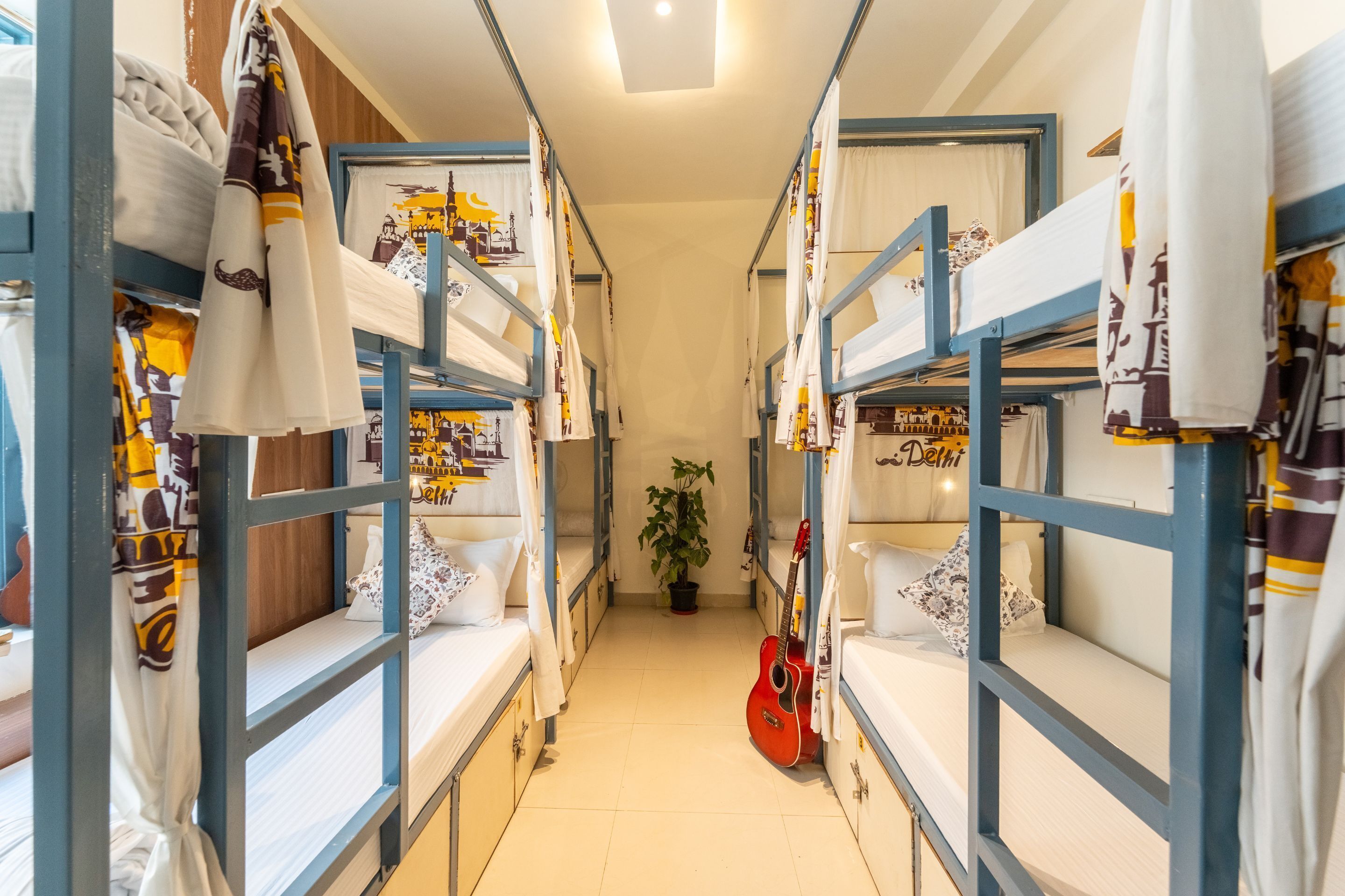 A narrow corridor with bunk beds on either side, a potted plant in the center, and a guitar leaning against one of the beds.