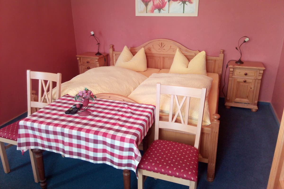 A bedroom with a checkered table, two chairs, a bed with yellow pillows, a nightstand with a lamp, and a painting of roses on the wall.