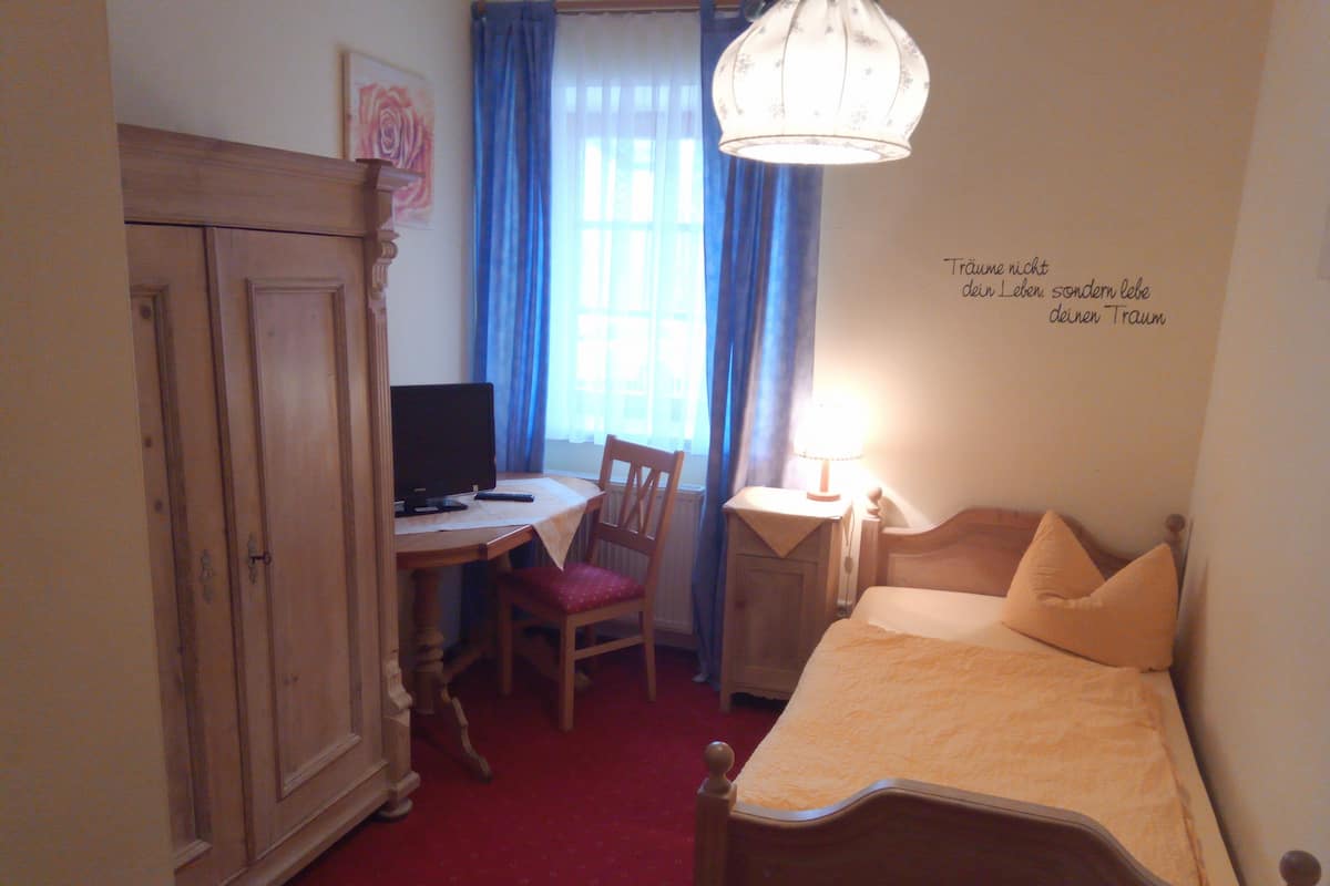 A bedroom with a bed, a desk with a computer, a chair, a wardrobe, and a window with curtains.