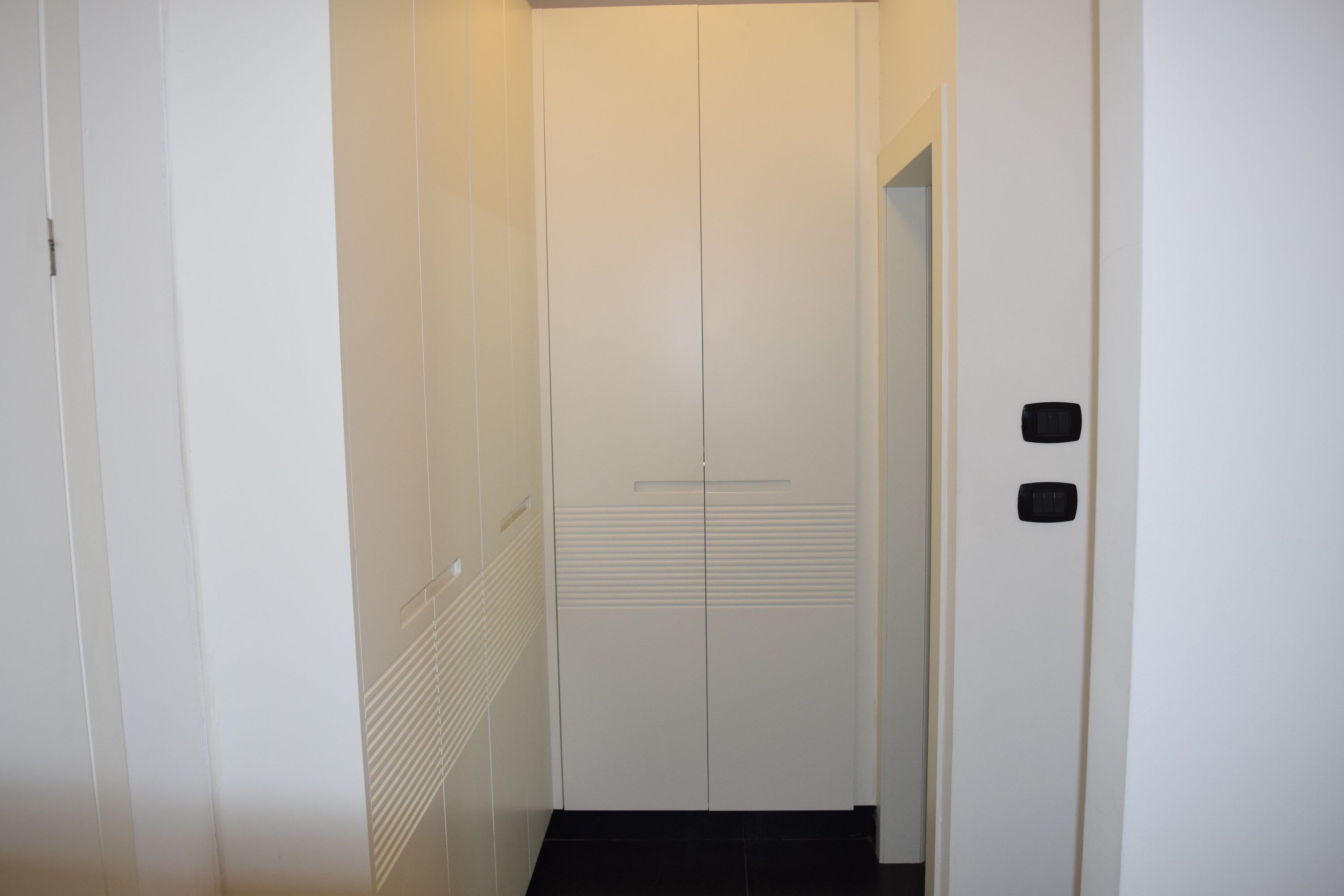 A narrow hallway with a white door at the end, flanked by white walls and a black tiled floor.