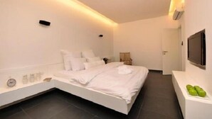 A modern bedroom with a large bed, bedside tables, a flat-screen TV, and a small seating area with a chair and a table. - Tal Kinneret (Emek HaYarden)