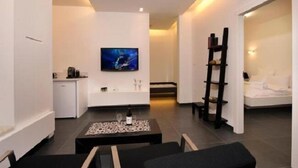 A modern hotel room with a kitchenette, a flat-screen TV mounted on the wall, a black coffee table with a bottle and glasses, and a bed with white bedding. - Tal Kinneret (Emek HaYarden)