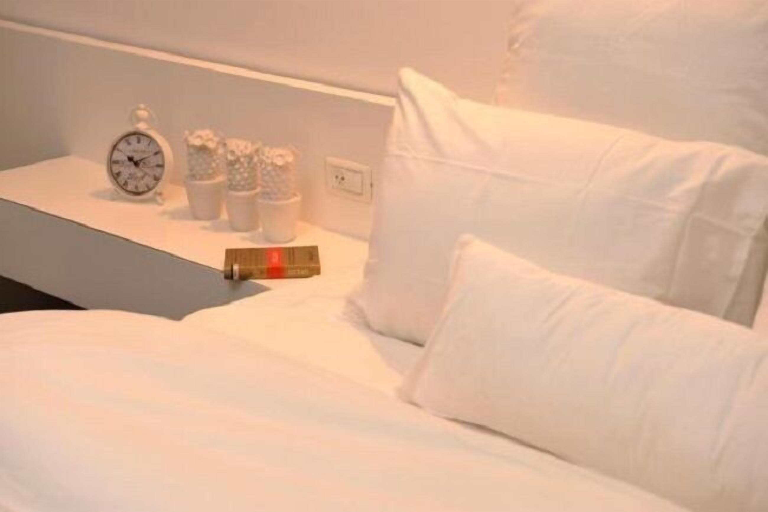 A neatly made bed with white linens and pillows, a bedside table with a clock and decorative items, and a power outlet on the wall.