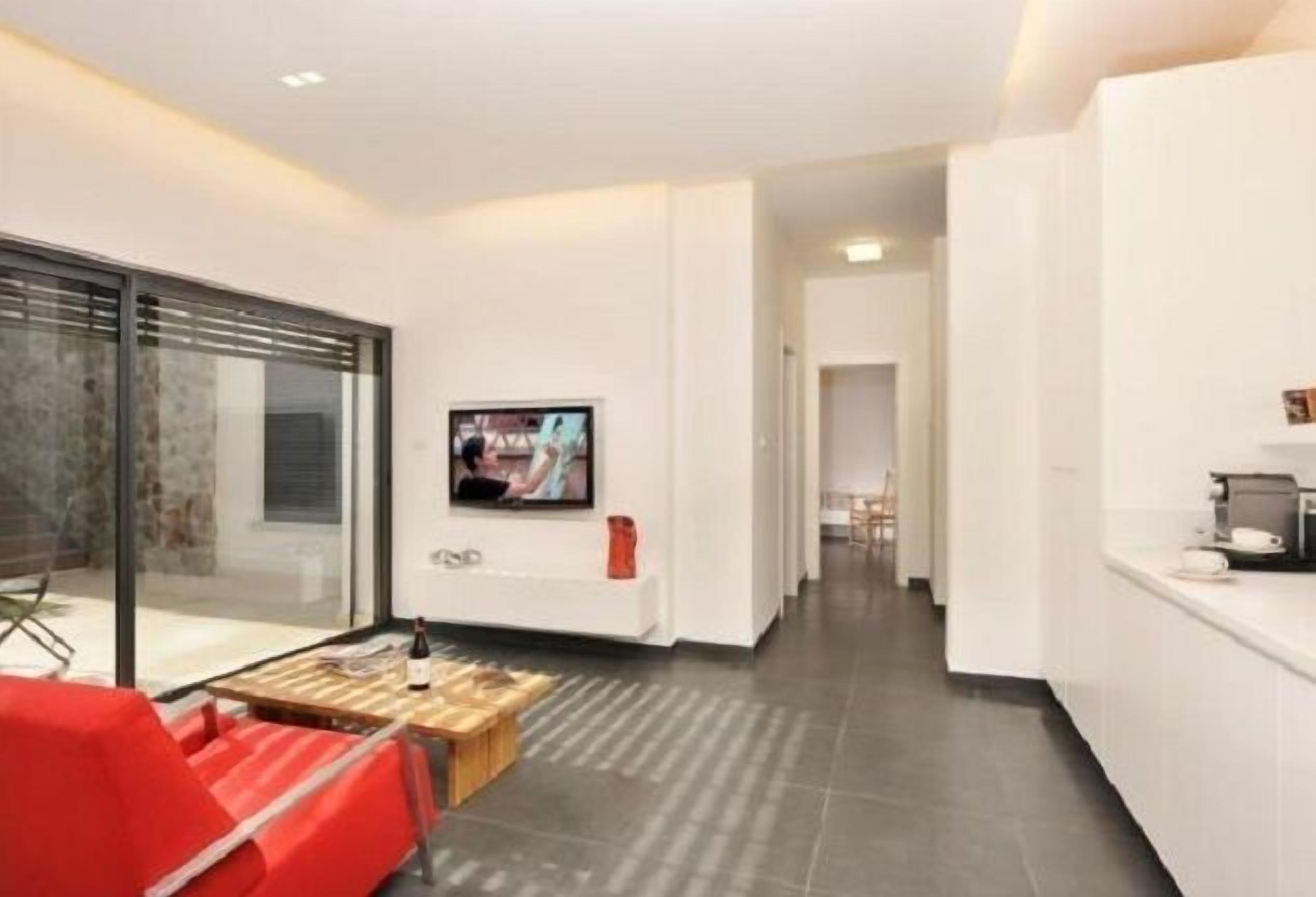A modern hotel room with a large sliding glass door, a flat-screen TV mounted on the wall, a wooden coffee table, and a red chair.